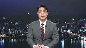 안경을 쓴 남성 뉴스 앵커가 정장을 입고 앉아 있으며 배경은 야간 도시 전경임.