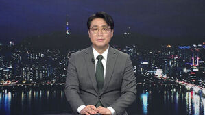 안경을 쓰고 정장을 입은 남성 앵커가 야경 배경의 뉴스 스튜디오에 앉아 있다.