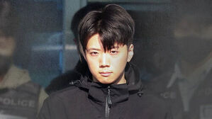 검은색 점퍼를 입은 젊은 남성이 경찰관들에게 둘러싸여 정면을 응시하는 모습.