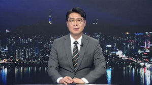 안경을 쓴 남성 뉴스 앵커가 정장을 입고 밤의 도시 야경을 배경으로 앉아 있다.