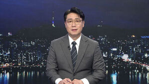안경을 쓴 남성 뉴스 앵커가 정장 차림으로 야경 배경의 스튜디오에 앉아 있다.